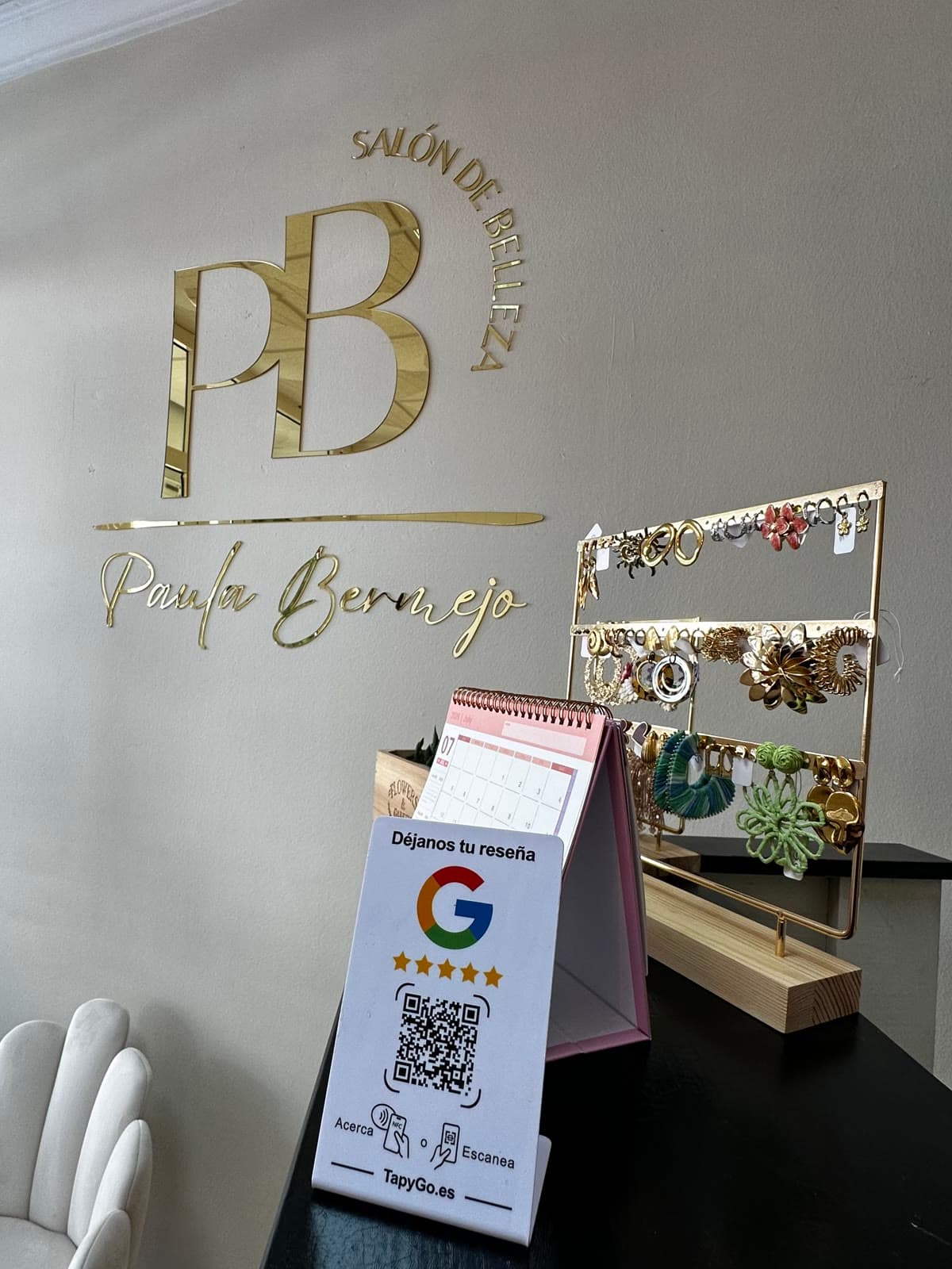 La placa TapyGo sobre el mostrador negro de un salón de estética, con el logotipo dorado del salón en la pared y un expositor de pendientes al lado.